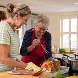 Waist-up shot of a senior lady with her daughter basting a fresh turkey fresh out of an oven.